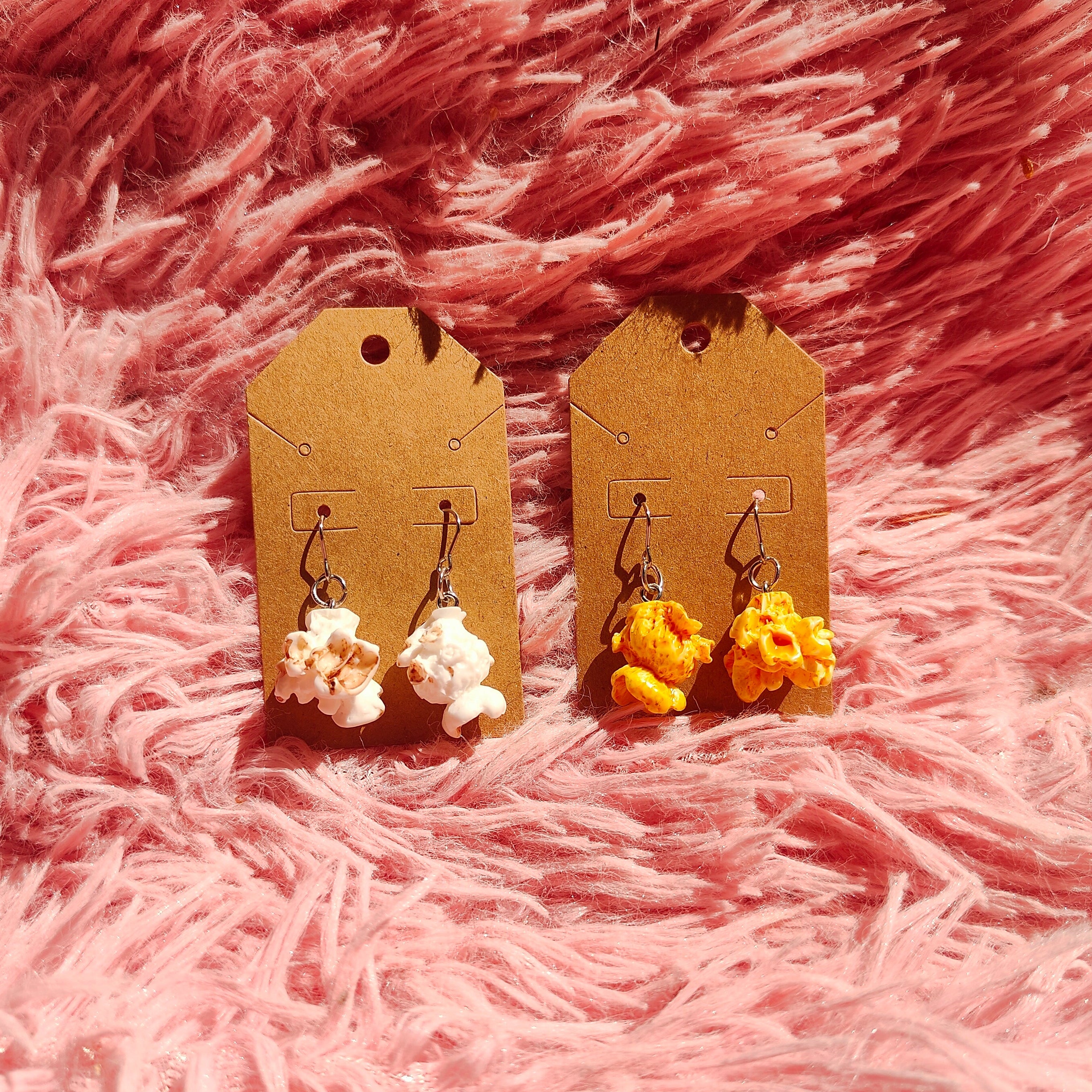 Two pairs of popcorn shaped earrings on display cards against a pink fluffy background