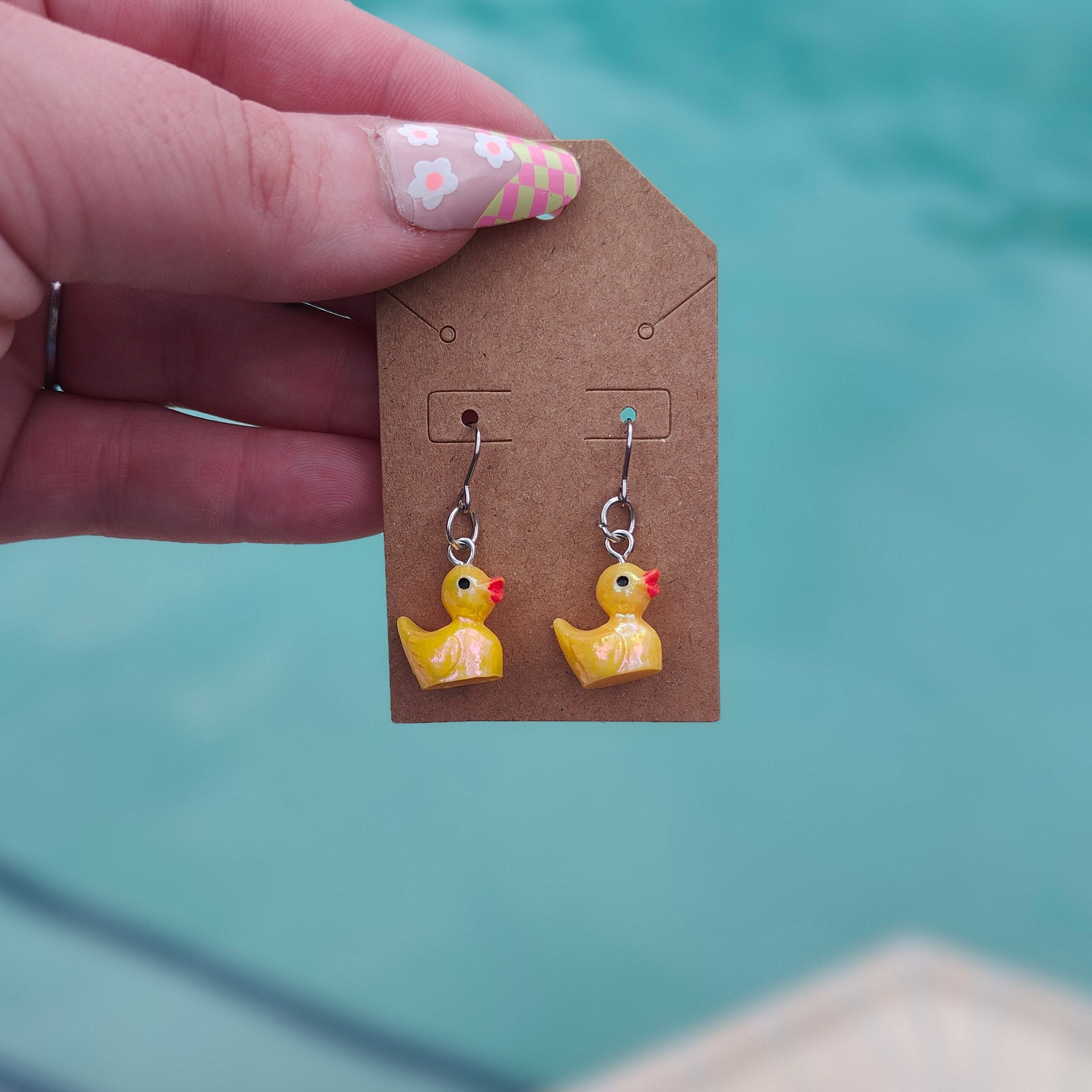 Hand holding a card with duck-shaped earrings against a pool background