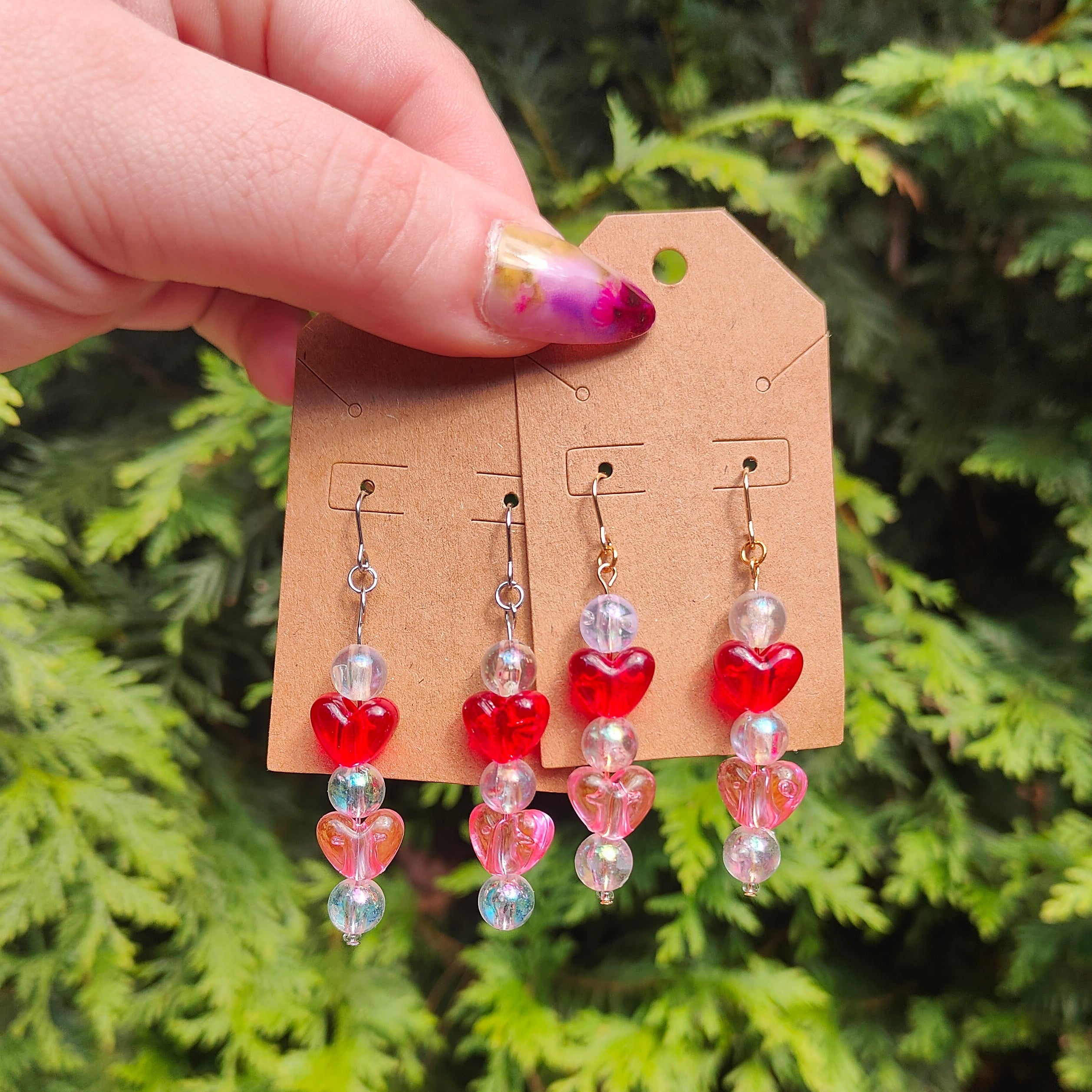 Hand holding a cardboard earring display with heart-shaped earrings against a green foliage background
