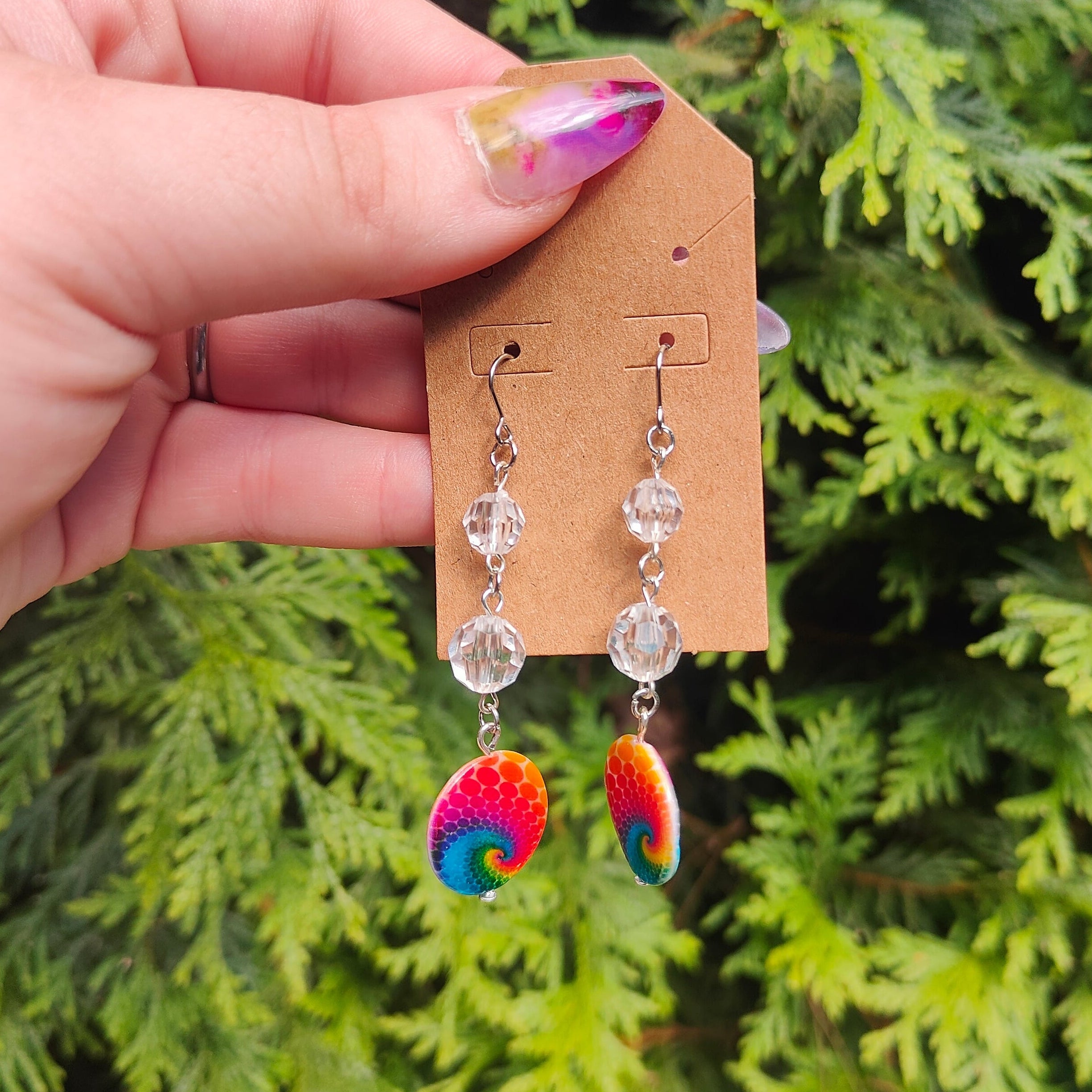 Hand holding a pair of earrings with a colorful design on a wooden card against a green leafy background.