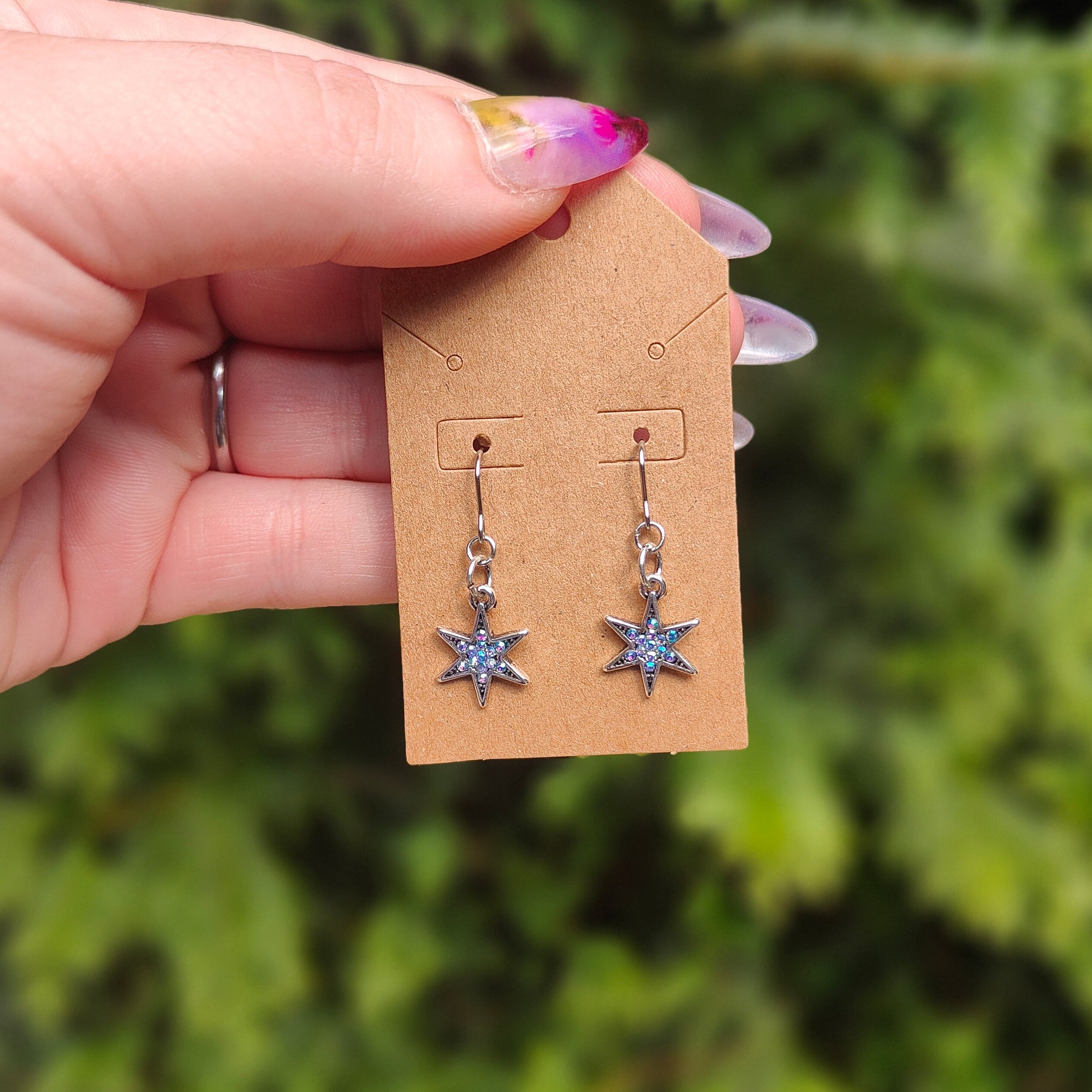 Hand holding a pair of star-shaped earrings on a cardboard display card against a green foliage background.