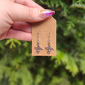 Hand holding a card with hummingbird earrings against a green foliage background