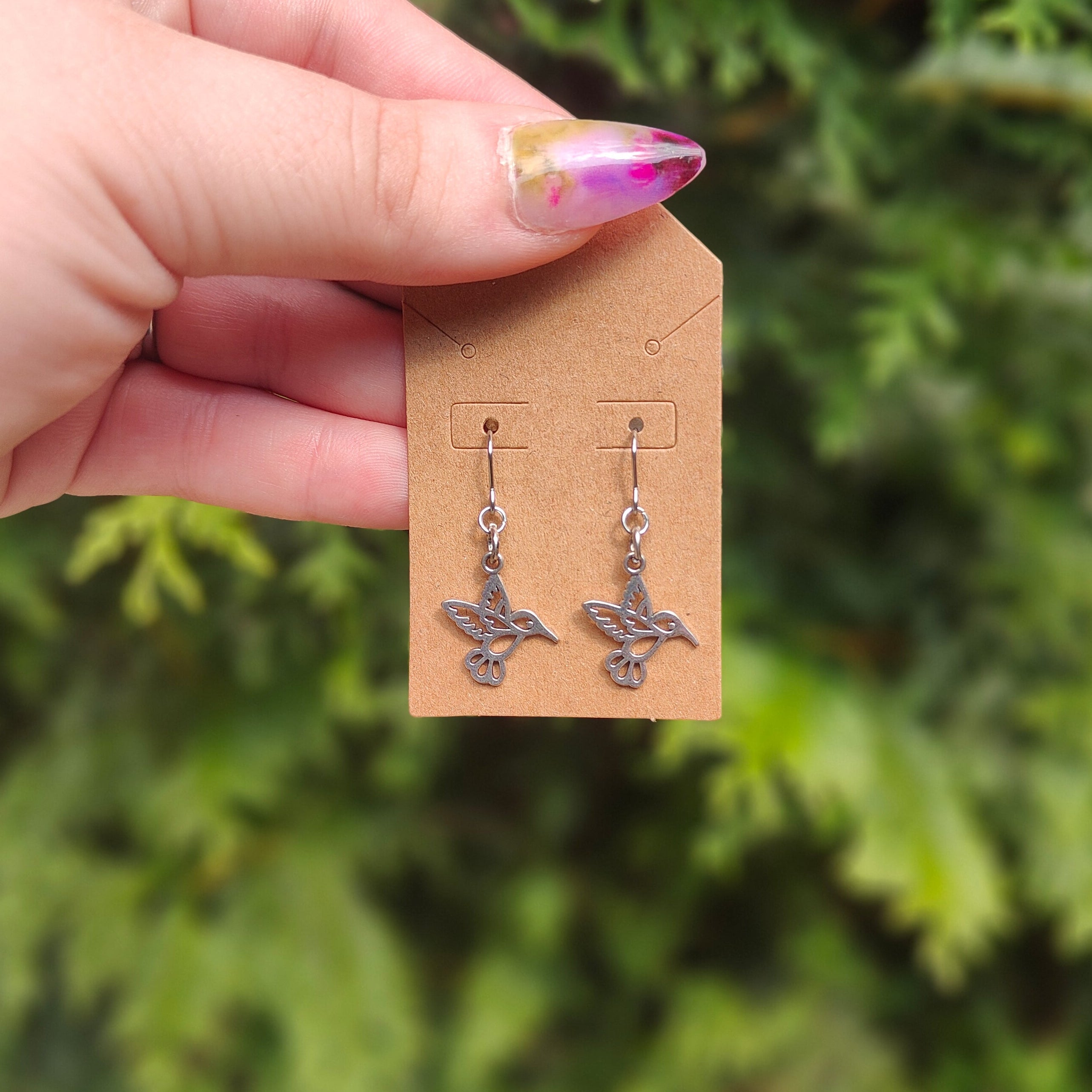 Hand holding a card with hummingbird earrings against a green foliage background